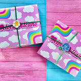 Two pastel pink boxes with white cloud pattern and wiggly pastel rainbow accents in the upper left and bottom right. Both boxes have a crochet flower tie wrapped around them. The boxes are on a pink and turquoise tinted wood background